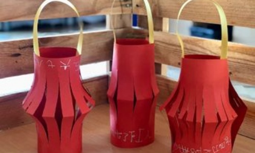 Three red chinese lanterns , made from card, placed on a table. The lanterns have gold paper handles and names written in mandarin characters.